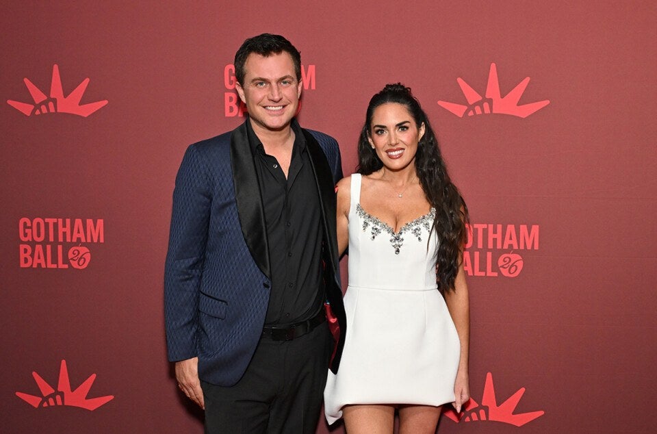 A man and a woman posing together in front of a red backdrop with "GOTHAM BALL 26" text and crown-like logos. The man is wearing a dark blue textured blazer with black lapels over a black shirt and black pants. The woman is wearing a short white dress with silver embellishments around the neckline and has long dark hair. Both are smiling.