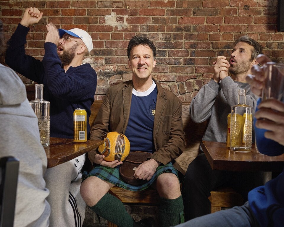 A man wearing a brown jacket, blue shirt, green tartan kilt, and green knee-high socks sits on a wooden bench against a brick wall, holding a yellow and brown soccer ball. Two men sit on either side of him; the man on the left wears a navy sweater and white cap, raising his fists excitedly, while the man on the right wears a gray sweatshirt and claps his hands. There are bottles and cans on the wooden tables in front of them.