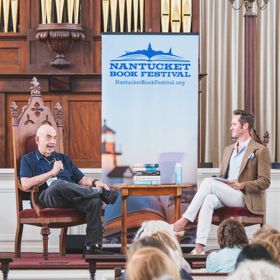 Two men seated on stage in wooden chairs, engaged in a discussion at the Nantucket Book Festival. A small wooden table between them holds a stack of books and a water bottle. Behind them is a large banner with the festival's name and website. The setting features wood paneling and organ pipes in the background. An audience is partially visible in the foreground.