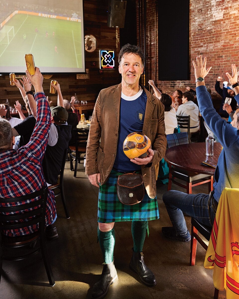 Man wearing a brown jacket, blue shirt, green and blue tartan kilt, green knee-high socks, and black boots, holding a vintage-style soccer ball in a lively pub setting with people cheering and raising drinks while watching a soccer game on a large screen.