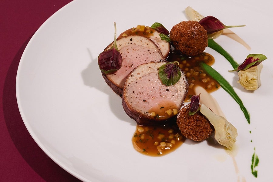 Two slices of roasted meat garnished with small green leaves, served with two round, breaded croquettes, artichoke hearts, and drizzled with a brown sauce and green puree on a white plate.