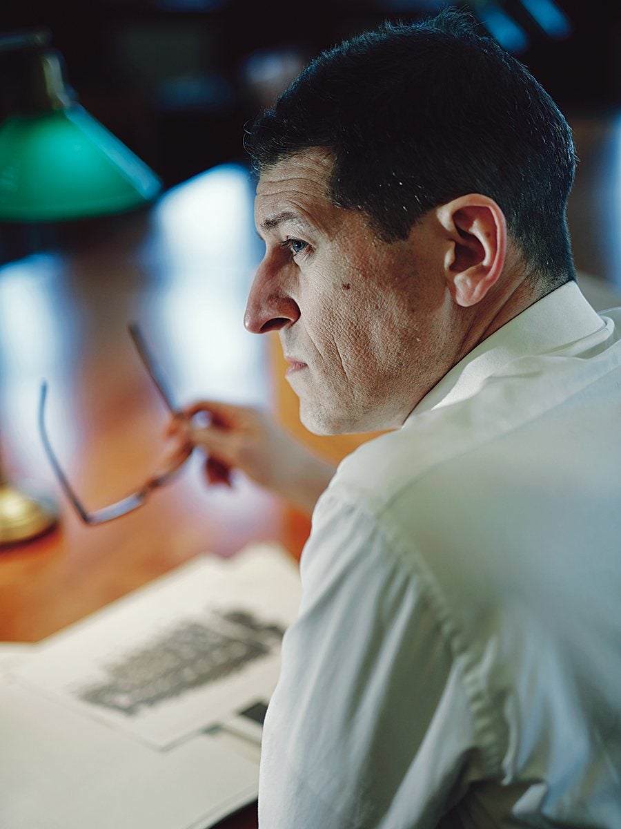Man with short dark hair wearing a white shirt, sitting at a wooden desk, holding a pair of glasses in one hand, looking thoughtfully to the side. An open book or document with black and white images is on the desk, and a green desk lamp is visible in the background.