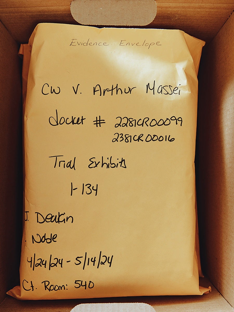 A large yellow evidence envelope inside a cardboard box. The envelope is labeled with handwritten text including "Evidence Envelope," "CW v. Arthur Massei," docket numbers "2281CR00099" and "2381CR00016," "Trial Exhibit 1-134," the name "J. Deakin," the word "Node," dates "4/24/24 - 5/14/24," and "Ct. Room: 540.