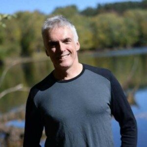 A middle-aged man with short gray hair is smiling and standing outdoors near a body of water. He is wearing a long-sleeve shirt with a dark gray front and black sleeves. The background features blurred trees and a clear blue sky.