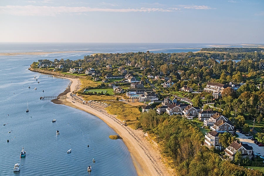 A coastal residential area with numerous houses surrounded by trees and greenery, bordered by a sandy beach and calm blue water with several small boats anchored nearby. The scene is captured under a clear sky with soft sunlight.