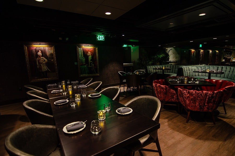 A dimly lit restaurant interior featuring a long dark wooden table set with plates, napkins, glasses, and small candle holders. Surrounding the table are cushioned chairs in muted tones. In the background, there are round tables with plush red velvet chairs and a curved light blue banquette. The walls are dark with framed portraits and illuminated exit signs above doorways. The floor is wooden, and the overall ambiance is cozy and intimate.