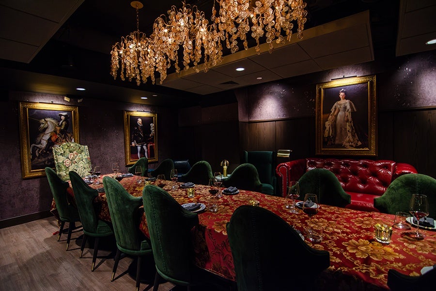 A dimly lit, elegant dining room featuring a long table covered with a red and gold floral tablecloth. The table is surrounded by green upholstered chairs and set with wine glasses and napkins. Above the table hangs a decorative chandelier with numerous amber-colored crystals. The walls are dark with wood paneling and adorned with three framed classical portraits. A red leather tufted sofa is positioned against one wall. The floor is light wood, contrasting with the darker tones of the room.