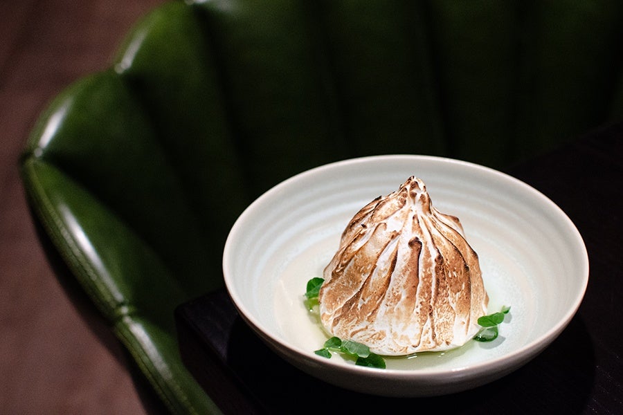 A toasted meringue dessert served in a white bowl, garnished with small green leaves, placed on a dark surface with a green upholstered chair in the background.