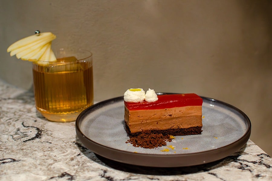 A slice of layered chocolate mousse cake with a red jelly top, garnished with two dollops of whipped cream, served on a gray plate with a dark rim. Next to the plate is a glass of amber-colored drink garnished with thinly sliced apple fanned out and secured with a small metal pick. The setting is on a marble countertop with a neutral background.