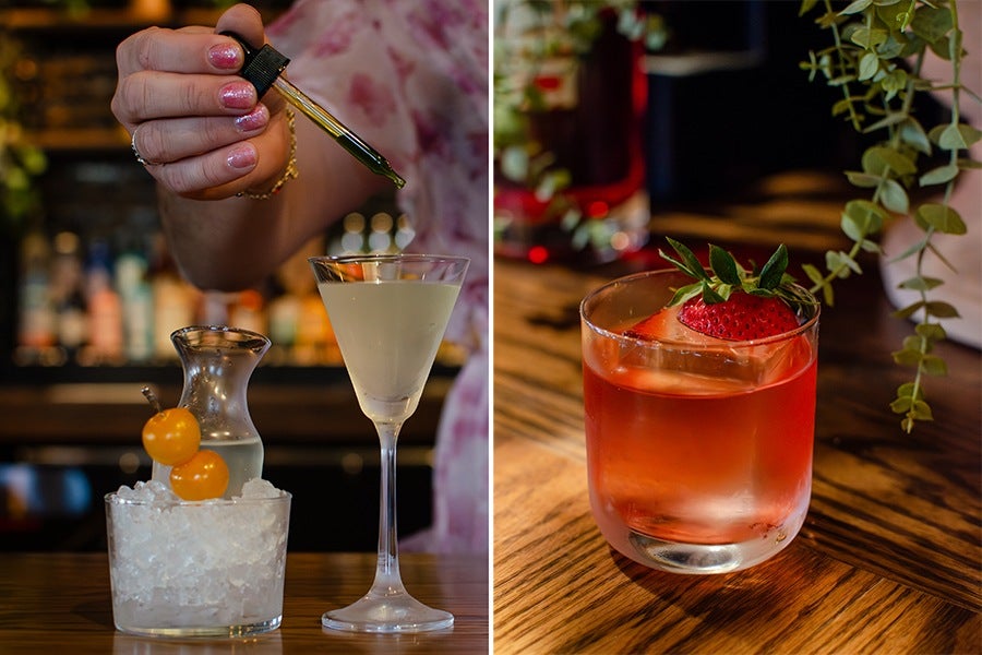 Left side: A hand with pink glittery nail polish is holding a dropper above a cocktail glass filled with a pale yellow drink. In front of the glass is a small container filled with crushed ice, two yellow cherry tomatoes on a skewer, and a small carafe with a clear liquid. The background is a blurred bar setting. Right side: A short glass with a reddish-pink cocktail, containing a large ice cube and garnished with a fresh strawberry and green leaves. The glass is on a wooden surface with some green foliage hanging nearby.