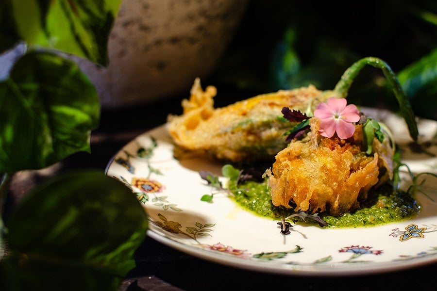 Two pieces of golden-brown fried stuffed squash blossoms served on a floral-patterned plate with a green herb sauce and garnished with a small pink edible flower and microgreens.