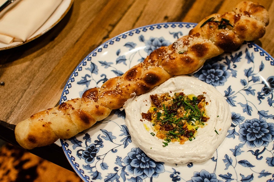 A twisted, golden-brown breadstick sprinkled with herbs and salt, served on a blue and white floral plate alongside a creamy white dip garnished with chopped herbs, nuts, and a drizzle of oil.