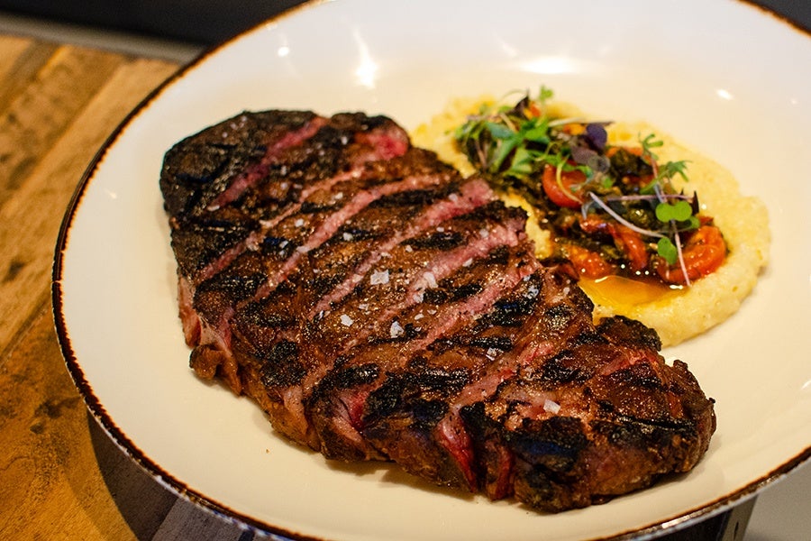 Grilled steak sliced and arranged on a white plate with a side of creamy polenta topped with roasted cherry tomatoes, herbs, and microgreens. The steak has visible grill marks and is seasoned with coarse salt.