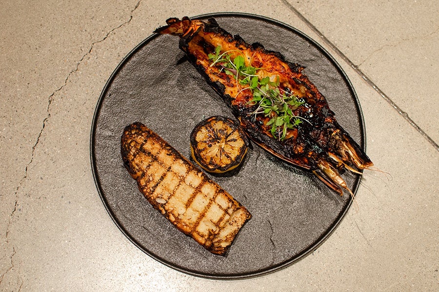 Grilled shrimp topped with microgreens, a charred lemon half, and a grilled piece of bread served on a round black plate.