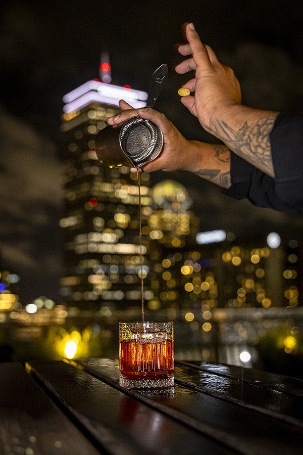 A hand with tattoos is pouring a dark liquid through a metal strainer into a glass filled with ice and an amber-colored drink, set on a wooden surface with a blurred cityscape and illuminated buildings in the background at night.
