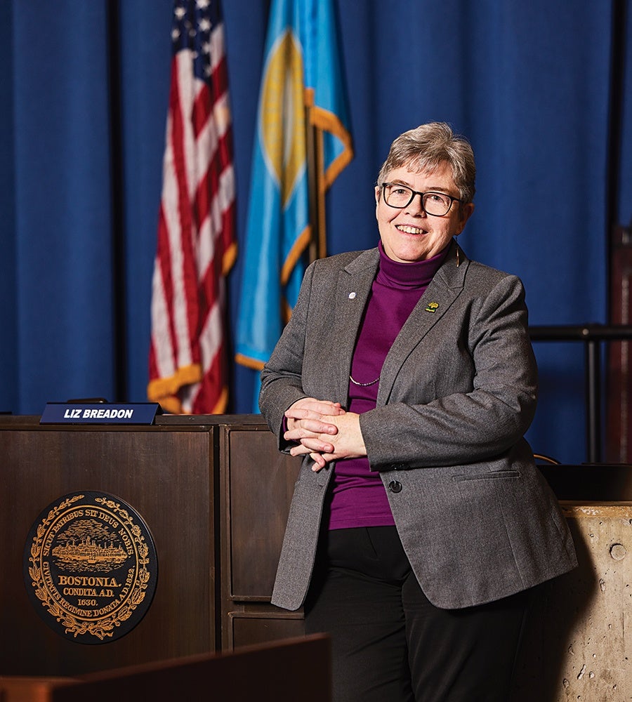 A person with short gray hair and glasses is smiling and leaning against a wooden podium with a Boston city seal and a nameplate that reads "Liz Breadon." They are wearing a gray blazer over a purple turtleneck and black pants. Behind them are two flags, one of the United States and another with blue and yellow colors, set against a blue curtain backdrop.
