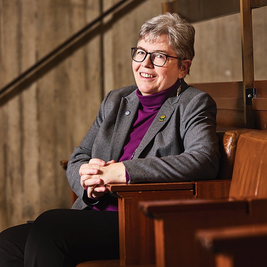 A middle-aged person with short gray hair and glasses is sitting in a wooden chair with brown leather cushions. They are wearing a gray blazer over a purple turtleneck and black pants, smiling warmly with hands clasped together resting on the armrest. The background features a textured, neutral-colored wall.