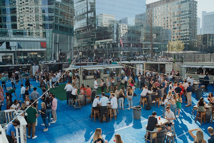Outdoor urban event with a large crowd of people socializing on a blue and green floor area surrounded by modern glass buildings. Several food or drink stalls are set up, and people are sitting on stools around barrel tables. String lights are hung overhead, adding to the festive atmosphere.