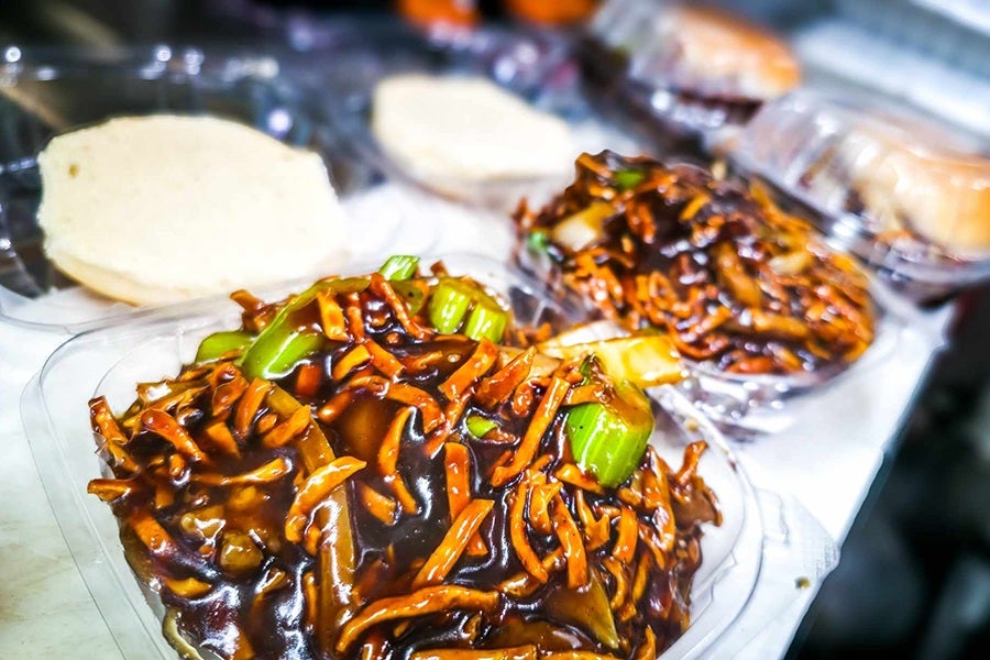 Open hamburger buns sit in clear plastic takeout containers, covered with chow mein noodles and celery in a thick, dark brown gravy.