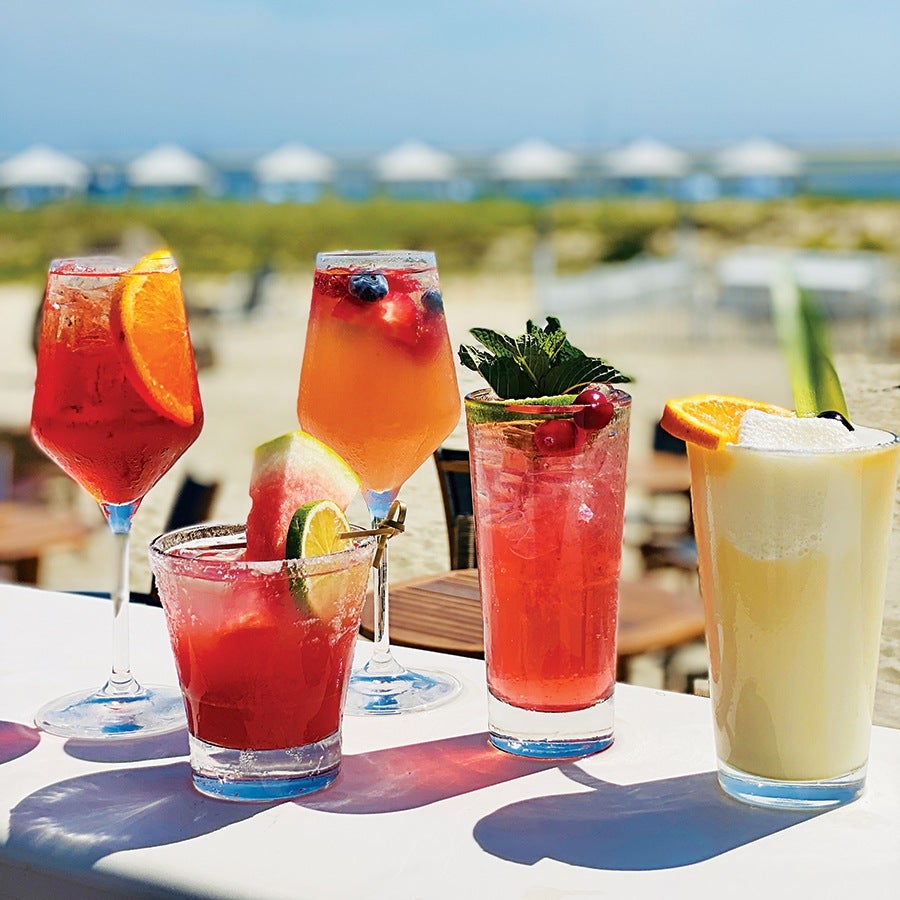 Five colorful cocktails are arranged on a white table outdoors, with a blurred beach and ocean background. The drinks include a red cocktail with an orange slice in a wine glass, a light orange cocktail with berries in a wine glass, a red cocktail with a watermelon and lime garnish in a short glass, a tall pink cocktail with mint and red berries, and a creamy yellow cocktail with an orange slice and celery stalk.