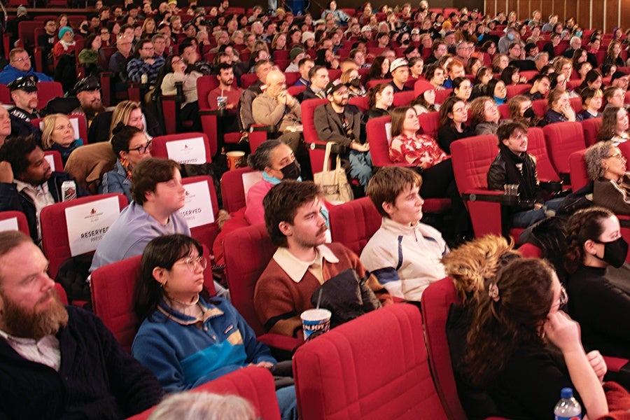 A large audience seated in a theater with red cushioned chairs, attentively watching an event. Some people are wearing masks, and a few seats have "Reserved" signs. The crowd is diverse, with individuals of various ages and styles.