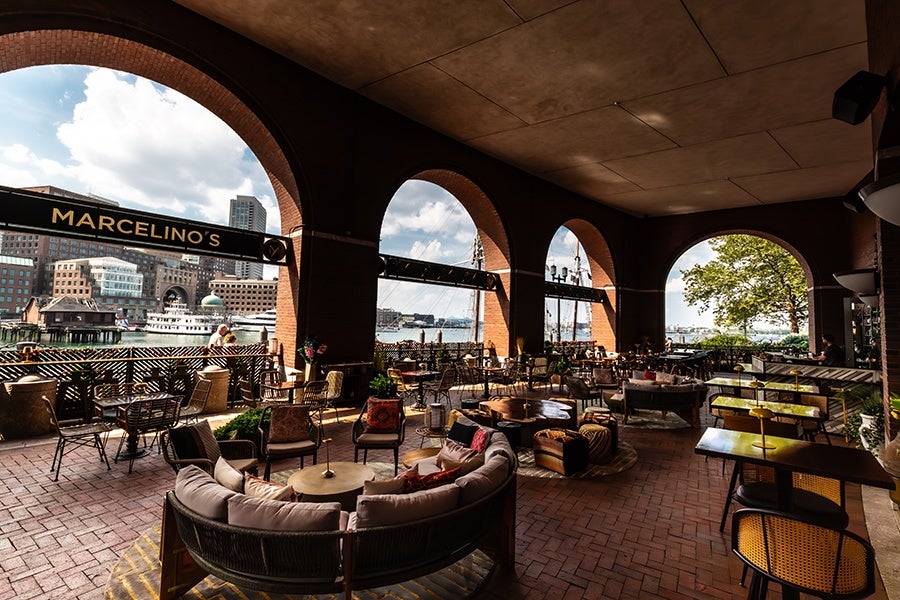Outdoor seating area of a restaurant named Marcelino's, featuring a variety of tables and chairs arranged under large brick archways. The space overlooks a waterfront with boats and city buildings visible in the background. The seating includes cushioned sofas, armchairs, and dining tables, with some greenery and decorative elements enhancing the ambiance.
