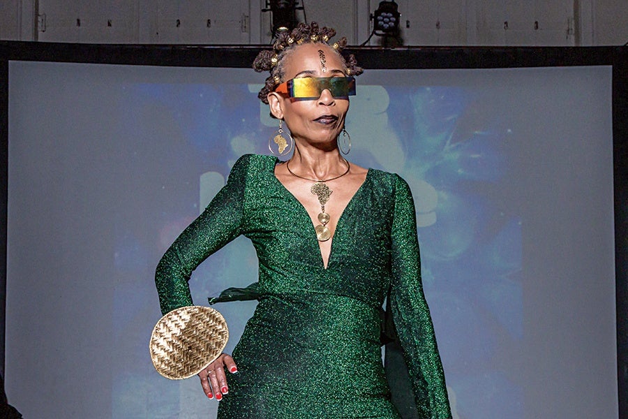 A person wearing a shimmering green dress with a deep V-neckline and long sleeves, accessorized with large gold earrings, a gold necklace, and futuristic, multicolored sunglasses. Their hair is styled in small, twisted buns adorned with gold beads, and they hold a round, woven gold clutch. The background features a blurred projection screen.