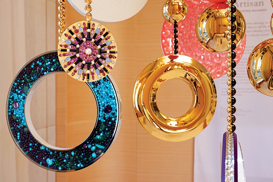 Decorative hanging ornaments featuring circular designs, including a large ring filled with small blue and green beads, a gold ring with a smooth reflective surface, and a smaller circular piece adorned with purple, white, and black beads arranged in a radial pattern. The ornaments are suspended by strings of gold and black beads against a wooden background.