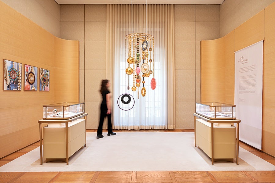 A modern, minimalist gallery space with light wood-paneled walls and a large white rug on a wooden floor. Two glass display cases with beige bases are positioned symmetrically on either side of the room, showcasing jewelry. A person dressed in black stands near the center, facing a large window covered with sheer white curtains. Hanging in front of the window is an artistic mobile featuring various colorful, circular jewelry pieces suspended from a gold frame. Three framed close-up photos of jewelry are mounted on the left wall, and a large text panel is displayed on the right wall.