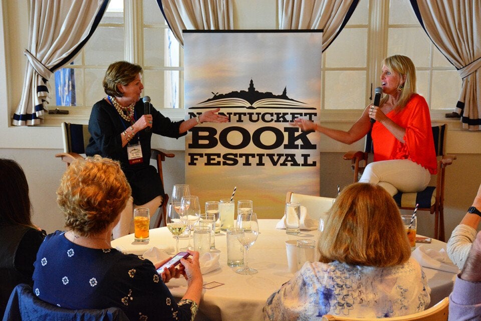 Two women are seated on stage chairs, each holding a microphone and engaged in conversation in front of a banner that reads "Nantucket Book Festival." The woman on the left wears a black outfit with a pearl necklace, while the woman on the right wears a bright orange top and white pants. In the foreground, several audience members sit around a round table with glasses and drinks. The setting appears to be a well-lit room with large windows and curtains.