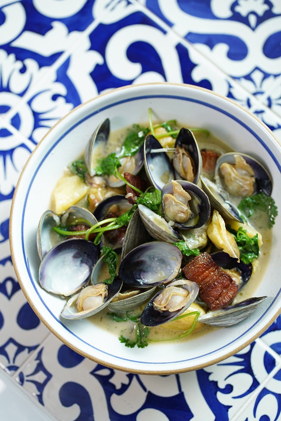 A bowl of soup featuring clams in their shells, garnished with fresh green herbs and pieces of crispy bacon, served in a white bowl with a blue rim on a blue and white patterned surface.