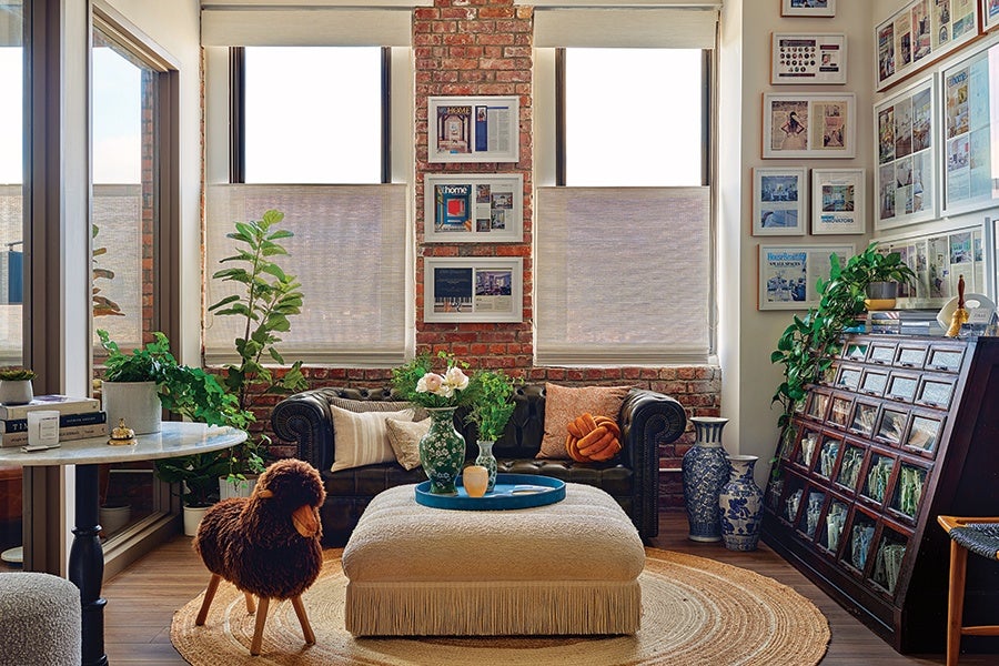 Cozy living room with a dark leather sofa adorned with various cushions, positioned against a brick wall with two large windows covered by light blinds. In front of the sofa is a beige ottoman with fringe, topped with a blue tray holding two green vases and a small yellow vase with flowers. A round woven rug lies beneath the ottoman. To the left, a small round table holds books and a candle, next to several green plants. On the right, a wooden cabinet with glass-fronted compartments displays framed pictures, with more plants on top. The walls are decorated with numerous framed photos and magazine covers. A plush dog-shaped stool stands on the rug near the ottoman.