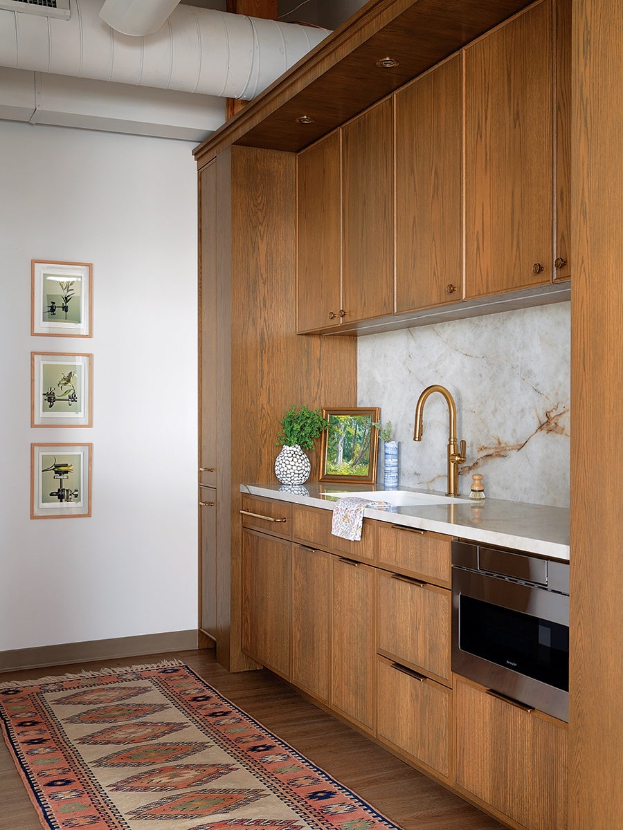 Kitchen area with wooden cabinets, a white marble backsplash, and a white countertop featuring a gold faucet. On the counter, there is a white vase with green foliage, a framed painting, a blue and white container, and a small brush. A built-in stainless steel microwave is integrated into the lower cabinets. The floor has a patterned rug with geometric designs in earthy tones. Three framed botanical prints hang on the adjacent white wall.