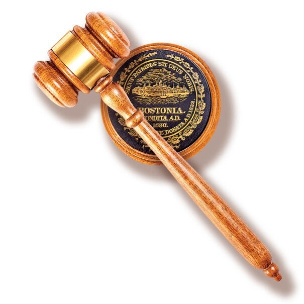 Wooden judge's gavel resting on a round wooden sound block with a black and gold emblem featuring the word "BOSTONIA" and other Latin text. The gavel has a polished finish with a gold metal band around the head.