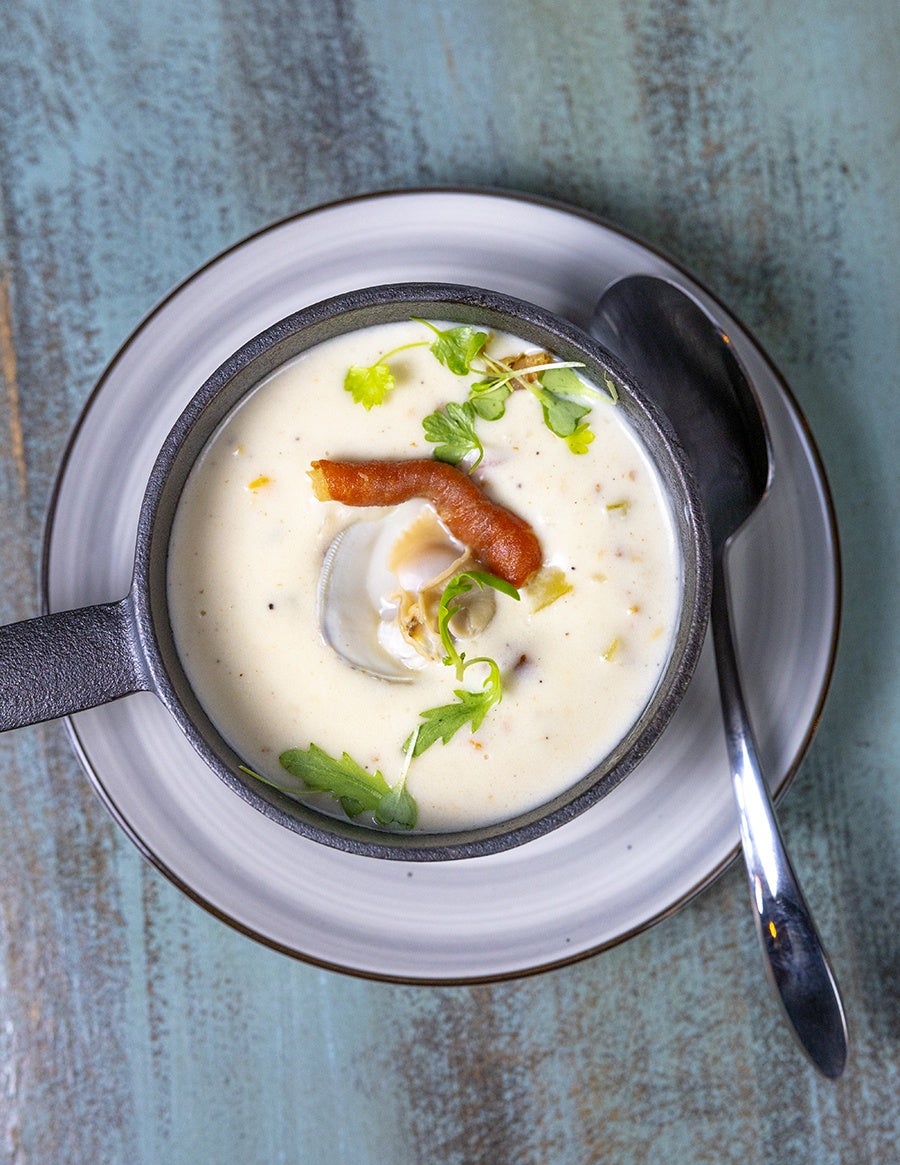 Creamy clam chowder served in a small black cast iron skillet, garnished with fresh green herbs and a piece of crispy bacon, placed on a white plate with a silver spoon beside it on a rustic blue wooden surface.