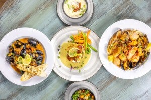 A top-down view of five plates of food arranged on a rustic wooden table. The central plate features a breaded fish fillet topped with lemon slices, served with green beans and carrot strips in a creamy sauce. To the left, a bowl of mussels in a tomato-based broth is garnished with lemon slices and accompanied by two pieces of toasted bread. To the right, a plate of seafood pasta includes clams, shrimp, and mussels in a light sauce with lemon wedges. Above the central plate, a small bowl of creamy soup garnished with herbs and a piece of bacon is visible. Below, a bowl of mixed vegetables including kale, carrots, and potatoes is present.