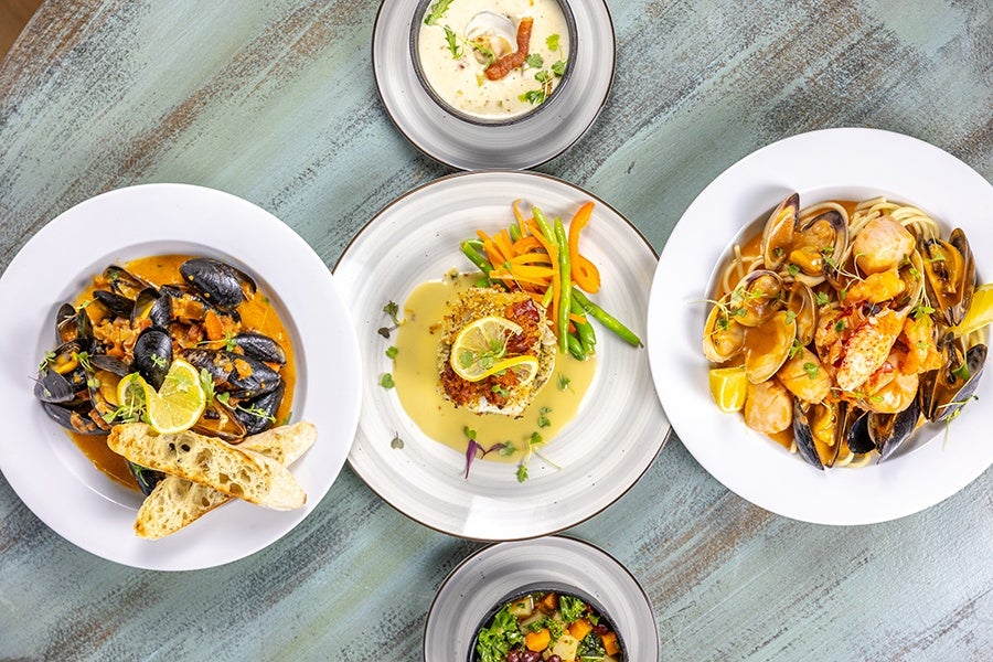 A top-down view of five plates of food arranged on a rustic wooden table. The central plate features a breaded fish fillet topped with lemon slices, served with green beans and carrot strips in a creamy sauce. To the left, a bowl of mussels in a tomato-based broth is garnished with lemon slices and accompanied by two pieces of toasted bread. To the right, a plate of seafood pasta includes clams, shrimp, and mussels in a light sauce with lemon wedges. Above the central plate, a small bowl of creamy soup garnished with herbs and a piece of bacon is visible. Below, a bowl of mixed vegetables including kale, carrots, and potatoes is present.