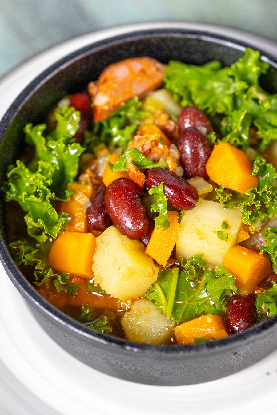 A bowl of hearty vegetable soup containing kale, kidney beans, diced potatoes, and chunks of orange squash or sweet potato, served in a black bowl on a white plate.