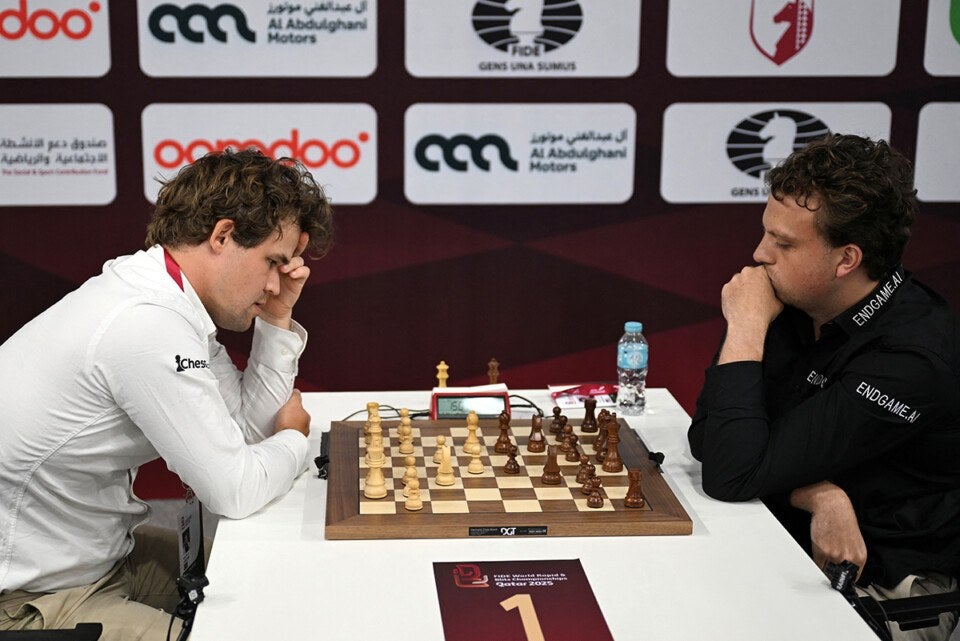 Two men are intensely focused on a chess game at a tournament table. The man on the left wears a white shirt with "chess.com" on the sleeve, while the man on the right wears a black shirt with "ENDGAME.AI" on the sleeves. The chessboard is set with wooden pieces, and a digital chess clock is positioned behind it. A water bottle and some papers are on the table. Behind them is a backdrop with various sponsor logos, including "Ooredoo," "Al Abdulghani Motors," and the FIDE logo. A sign on the table reads "FIDE World Rapid & Blitz Championships Qatar 2025" with the number 1.