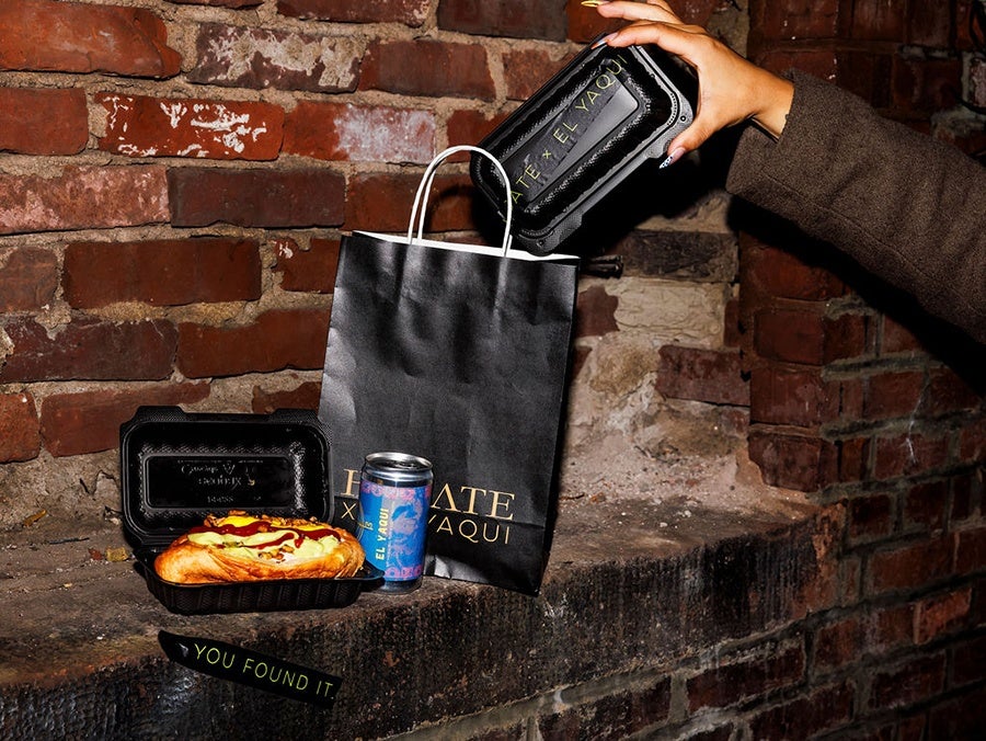 A hand is placing a black takeout container with gold lettering into a black paper bag with white handles, also featuring gold lettering. Next to the bag is an open black takeout container holding a sandwich topped with ketchup and mustard, and a blue and pink can with text. The items are set on a rough brick ledge against a brick wall. A small black label with green text reads "YOU FOUND IT.