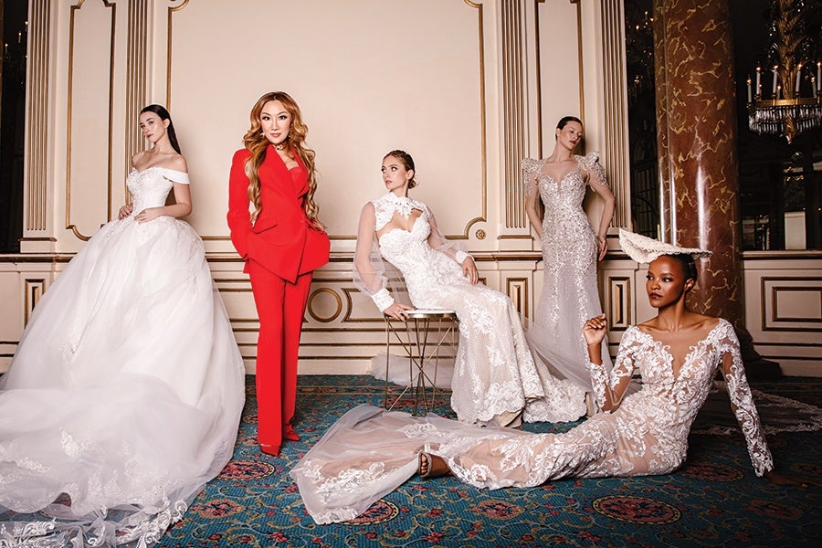 Five women are posing in an elegant room with ornate gold-trimmed walls and a patterned carpet. Four women wear intricate white bridal gowns with lace and sheer details, each with unique designs including off-shoulder, high neck, and embellished sleeves. One woman stands out in the center wearing a bold red pantsuit with a tailored blazer. The woman seated on the floor wears a long-sleeved lace gown and a matching lace headpiece. The overall scene conveys a high-fashion bridal theme.
