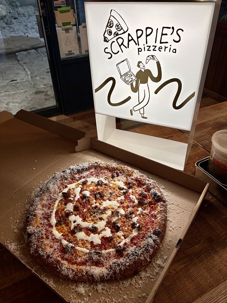 A pizza in an open cardboard box topped with tomato sauce, melted cheese, and a generous amount of grated cheese sprinkled around the edges. Behind the pizza is a lighted sign with the text "SCRAPPIE'S pizzeria" and a drawing of a person holding a pizza box in one hand and a game controller in the other. The scene is set on a wooden table near a door with snow visible outside.