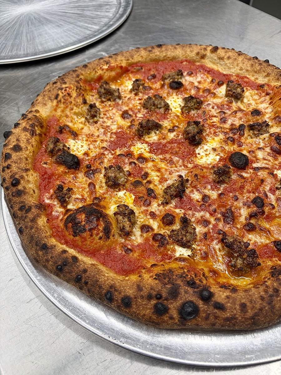 Pizza with a thick, well-charred crust topped with tomato sauce, melted cheese, and small chunks of browned sausage, served on a metal tray.