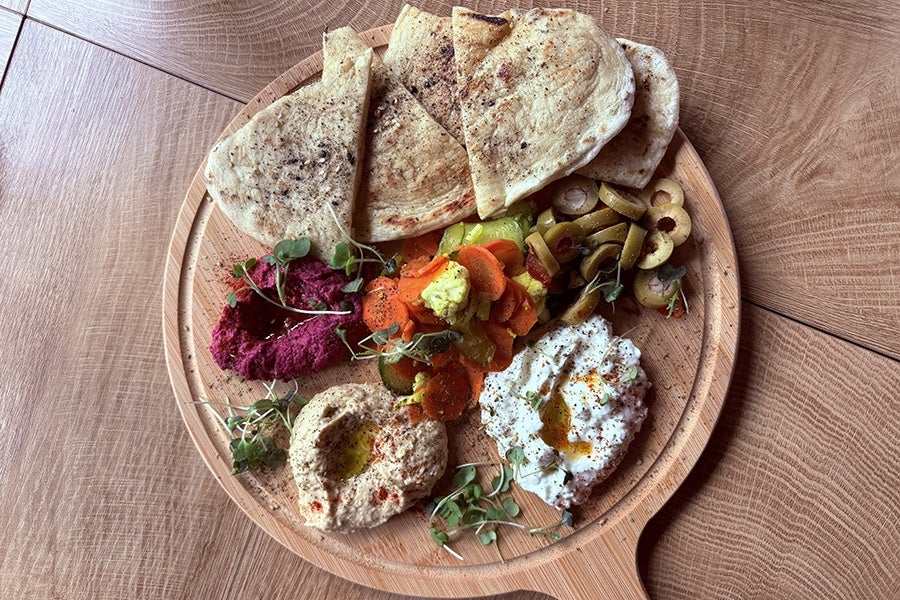 A wooden serving board with several pieces of seasoned pita bread, three types of dips (one pink beet-based, one beige hummus, and one white with olive oil), sliced green olives, pickled vegetables including carrots and cauliflower, and garnished with microgreens.