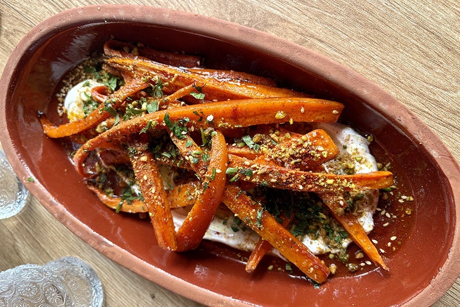 Roasted carrot sticks seasoned with spices and herbs, served on a bed of creamy white sauce in a rustic oval terracotta dish. The dish is garnished with chopped green herbs and crushed nuts or seeds.