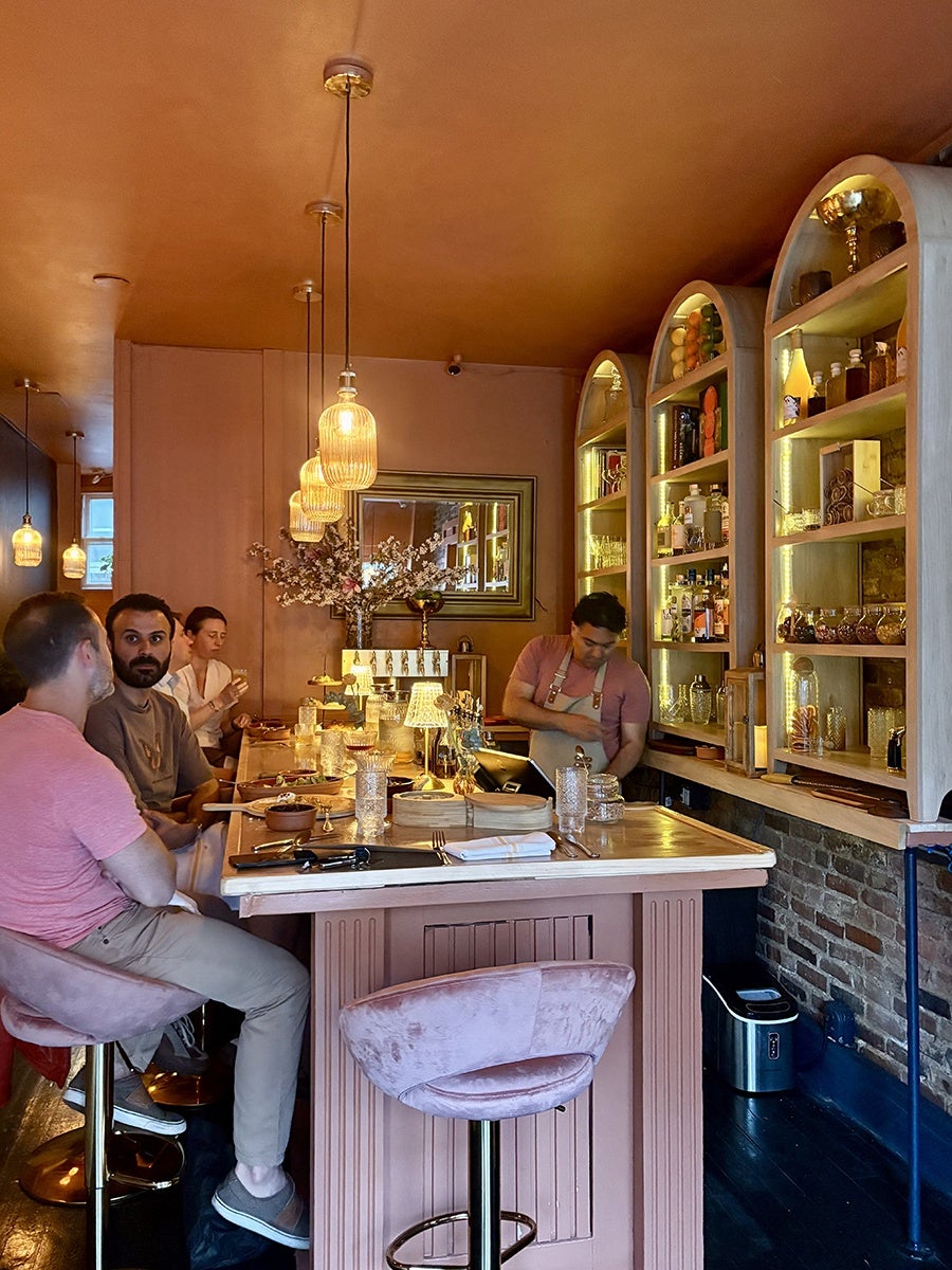 A cozy bar interior with warm orange lighting and a matching ceiling. Three people are seated on pink velvet bar stools at a wooden bar counter, which is adorned with glassware, plates, and decorative items. Behind the bar, a bartender in a pink shirt and apron is working. The back wall features arched shelves filled with bottles, glassware, and decorative objects, illuminated by soft yellow lights. A large mirror hangs on the wall, reflecting part of the bar area. The overall atmosphere is intimate and inviting.