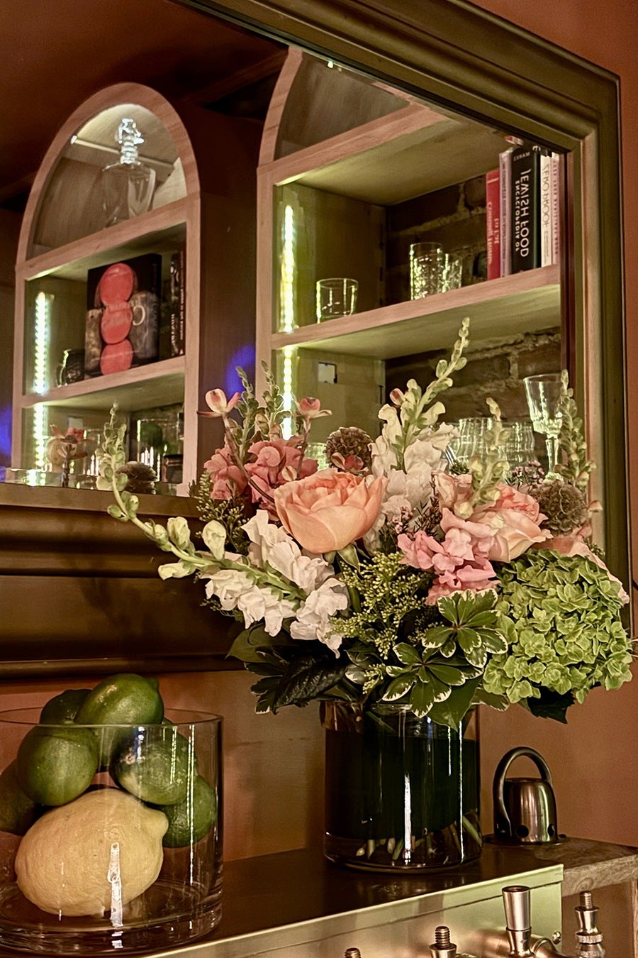A bouquet of mixed flowers including pink roses, white snapdragons, and green hydrangeas in a clear glass vase filled with water. The vase is placed on a wooden surface next to a glass container holding several green limes and one yellow lemon. In the background, there is a mirror reflecting a wooden shelf with books, glassware, and decorative items. The setting has warm lighting and a cozy ambiance.