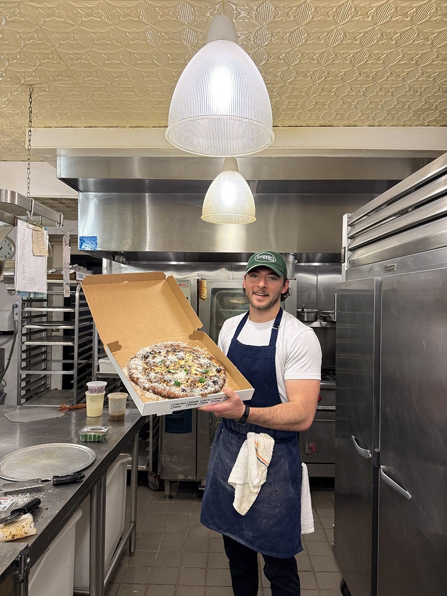 A man wearing a green cap, white t-shirt, and blue apron stands in a commercial kitchen holding an open pizza box with a pizza inside. The kitchen has stainless steel appliances, a tiled floor, and two hanging pendant lights above. A towel is tucked into the man's apron. Various kitchen items, including containers and utensils, are on the counter to the left.