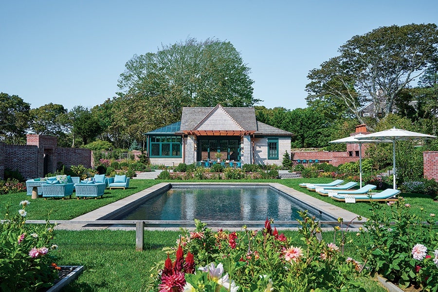A backyard scene featuring a rectangular swimming pool surrounded by a concrete border. On the left side of the pool, there is a seating area with light blue outdoor furniture including chairs and a sofa. On the right side, there are four light blue lounge chairs under two large white umbrellas. The pool area is bordered by well-maintained green grass and colorful flower beds in the foreground. In the background, there is a small house with a wooden pergola over a patio, surrounded by trees and brick walls. The sky is clear and blue.