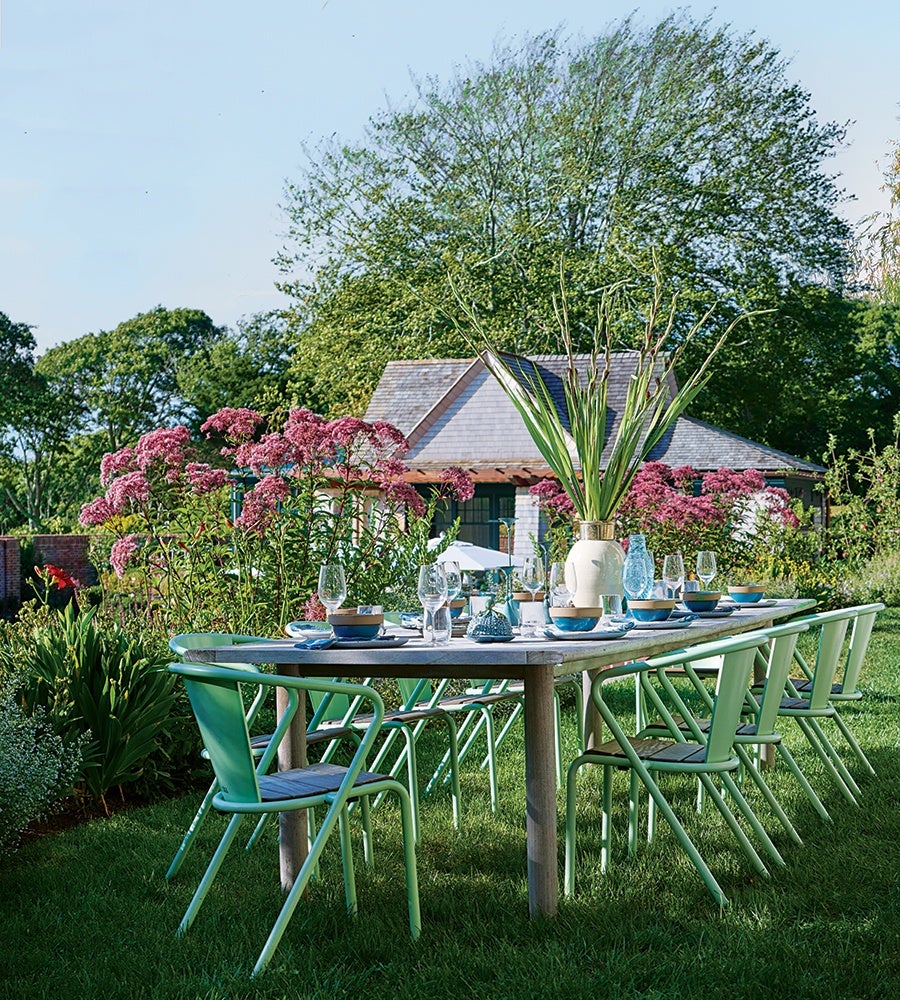 Long outdoor dining table set on grass with eight green chairs, decorated with glassware, bowls, and a large vase with tall green plants, surrounded by pink flowering bushes and trees, with a house in the background.